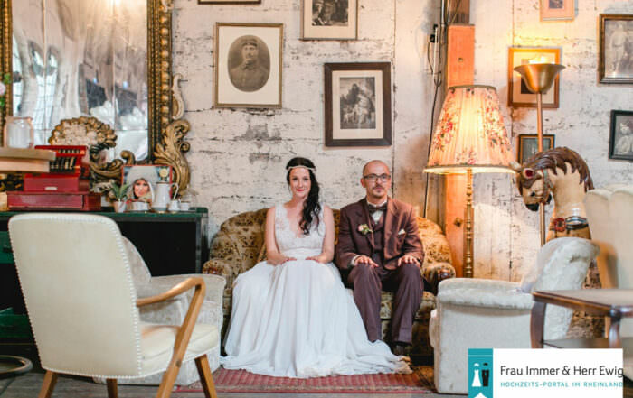 Hochzeitspaar sitzt auf Vintage Couch vor einer Wand mit Bilderrahmen