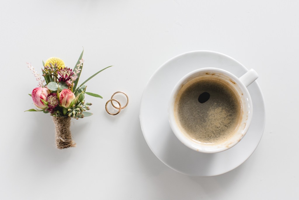 Hochzeitsfoto Idee: Blumenanstecker und Trauringe liegen neben einer Tasse mit Kaffee