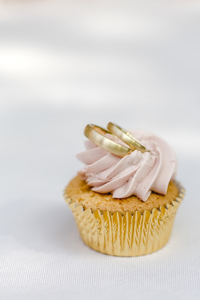 die goldenen Trauringe liegen auf einem Cupcake mit rosafarbener Creme