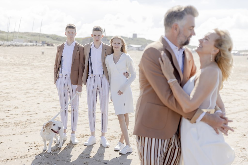 während die Eltern sich vorne umarmen, stehen die drei Kinder beim Familienfoto am Strand mit dem Hund im Hintergrund