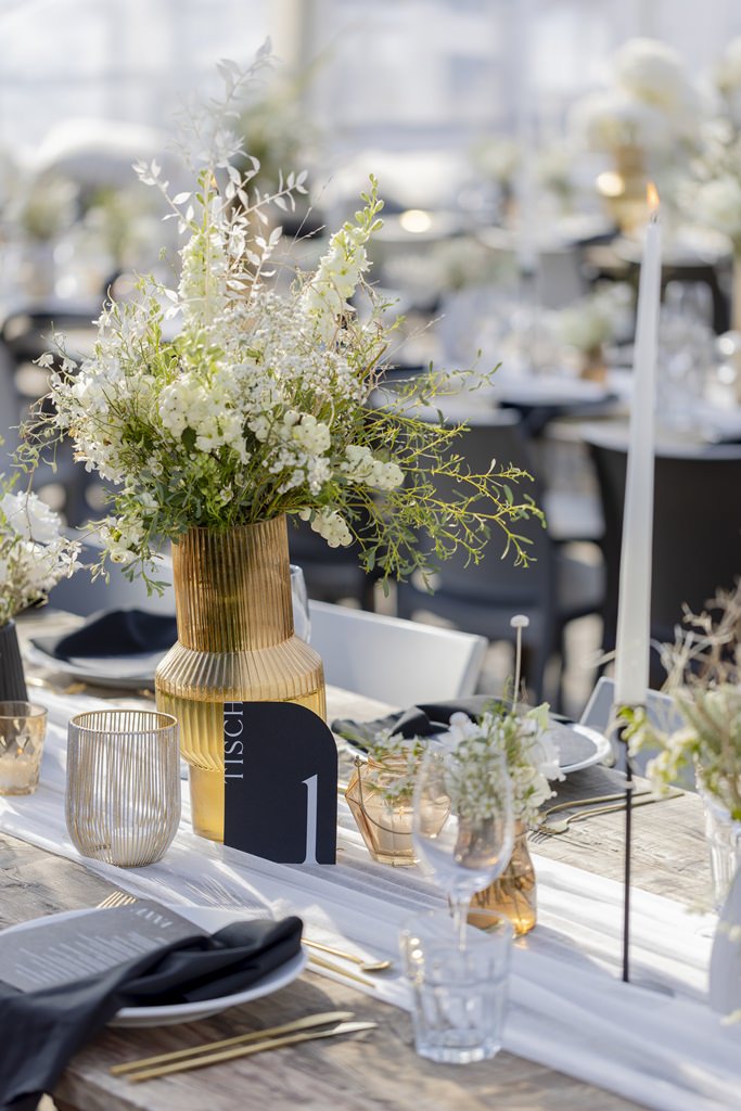 Hochzeitstischdeko mit goldener Vase, weißen Blumen, weißem Tischläufer, schwarzem Tischnummernschild und weißen Kerzen | Foto: Hanna Witte