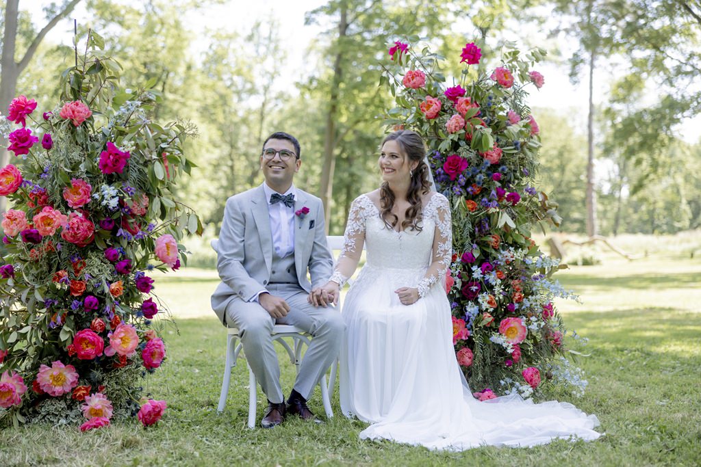 das Brautpaar sitzt während der outdoor Trauung zwischen opulenter Blumendeko in pink und blau | Hochzeitsfoto: Hanna Witte