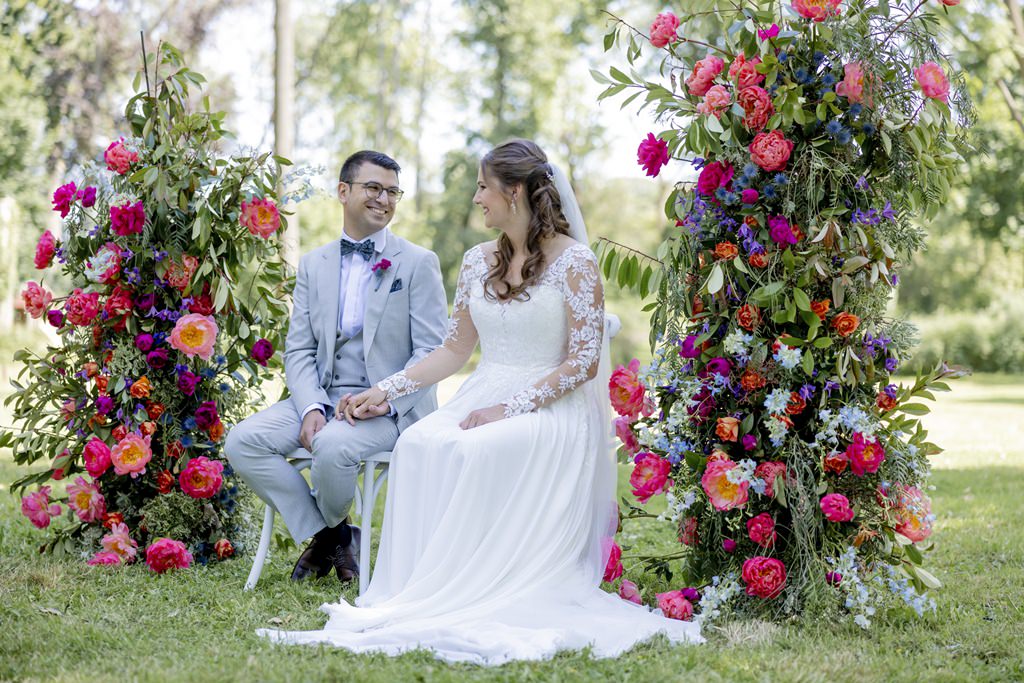 das Brautpaar sitzt während der Trauung Hand in Hand zwischen zwei Blumensäulen in pink und blau | Hochzeitsfoto: Hanna Witte