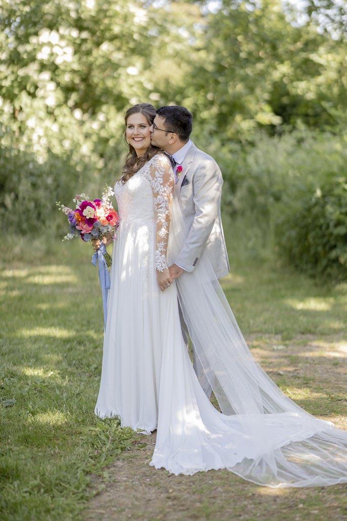 Paarfotopose: Der Bräutigam steht hinter der Braut und küsst sie zärtlich auf die Wange | Hochzeitsfoto: Hanna Witte
