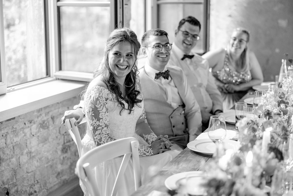 das Brautpaar sitzt während der Feier am gedeckten Hochzeitstisch und lacht | Hochzeitsfoto: Hanna Witte